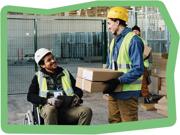 Happy young engineer in wheelchair looking at colleague carrying stack of packed cardboard boxes while talking to him during work in warehouse