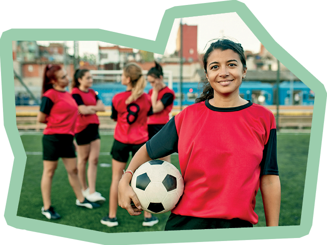 Close-up of smiling female Buenos Aires footballer in late 30s holding ball and looking at camera while teammates stand and talk in background.