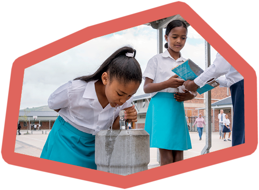Schoolgirl in uniform drinking water from a drinking fountain in school campus with classmates standing behind