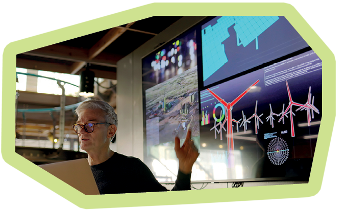 Stock photograph of a man conducting a seminar / lecture with the aid of a large screen. The screen is displaying data & designs concerning low carbon electricity production with solar panels & wind turbines. These are juxtaposed with images of conventional fossil fuel oil production.